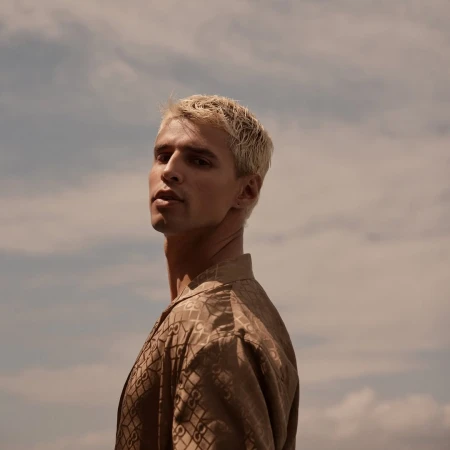 Blonde male model poses outdoors against cloudy sky wearing patterned button-down shirt in a low-angle portrait capturing fashion and style.