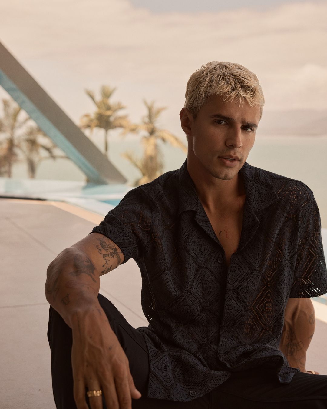 A blond male model poses in a dark patterned shirt and black pants outdoors with a pool, palm trees, and blue sky in the background.