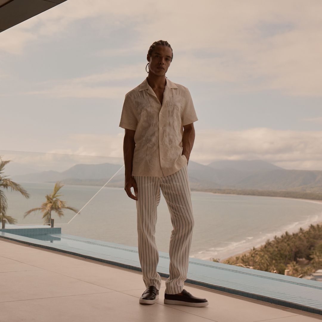 A full shot of a single adult male in a casual resort outfit with a serene coastal backdrop featuring the ocean and mountains.