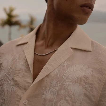 Close-up of a man wearing a cream embroidered short-sleeve shirt and a silver chain necklace against an outdoor coastal background.