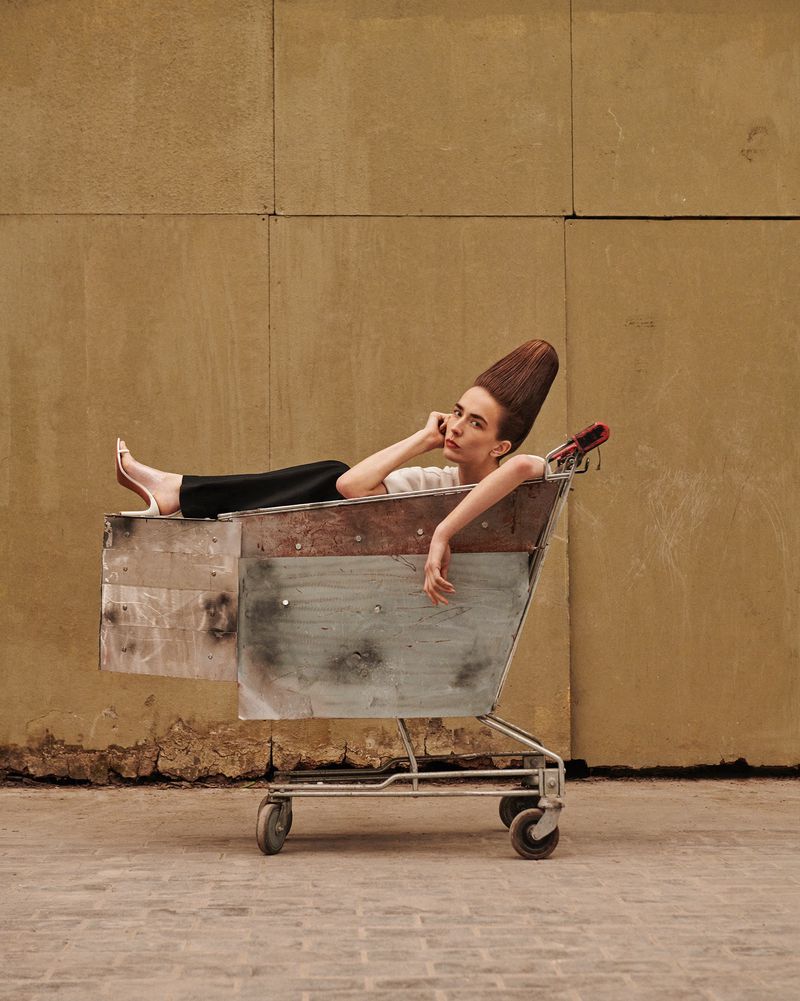 Woman with unique hairstyle rests in weathered shopping cart against neutral wall. Street fashion concept, posed shot, medium framing.