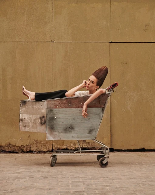 Woman with unique hairstyle rests in weathered shopping cart against neutral wall. Street fashion concept, posed shot, medium framing.