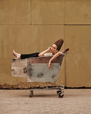 Woman with unique hairstyle rests in weathered shopping cart against neutral wall. Street fashion concept, posed shot, medium framing.