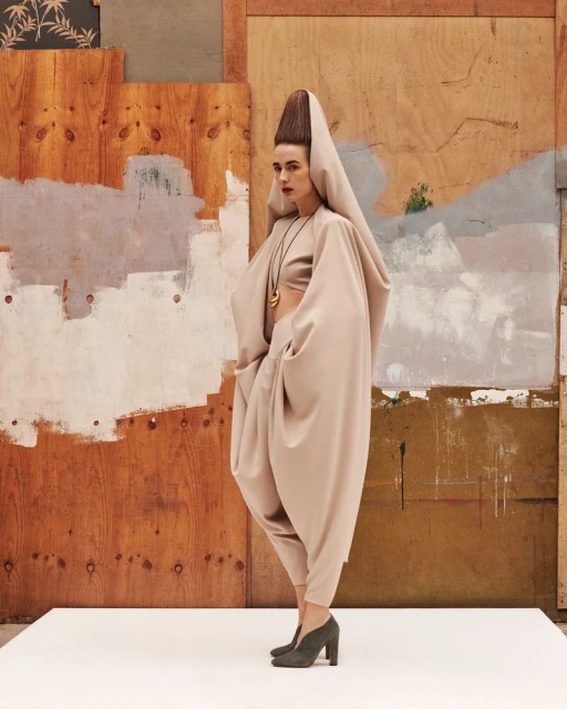 Woman in a dramatic, earth-toned draped couture outfit with matching heels and a tall hairstyle posing in front of a neutral backdrop.
