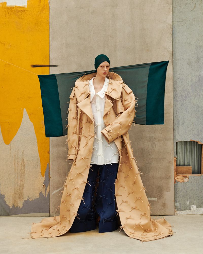Fashion editorial shot of a fair-skinned woman wearing an elaborate tan trench coat and a green cap against a textured background.