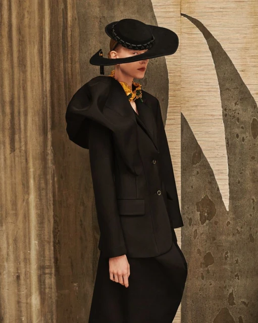 Fashion shot: a model in a black suit, wide-brimmed hat, and gold collar poses against a mottled brown and cream studio backdrop.
