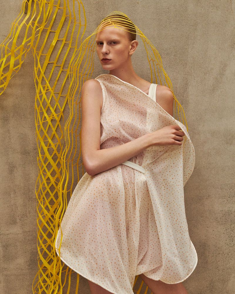 Woman in light pink dress with spotted pattern in front of a brown wall with yellow decorative structure, fashion editorial photography.