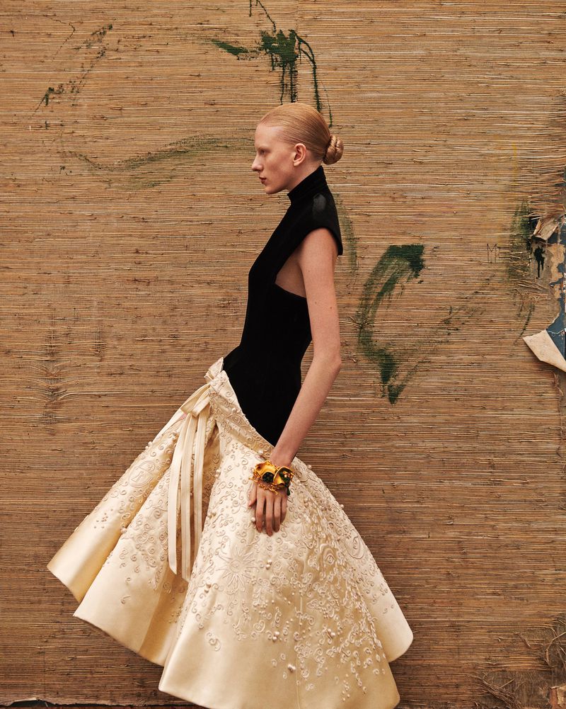 Elegant fashion portrait of a fair-skinned woman in a black top and light-colored gown, posing against a textured wall with paint.