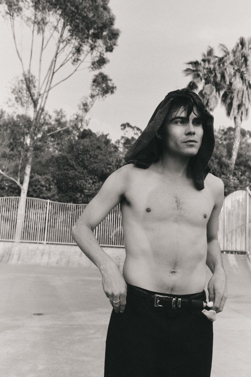 Black and white portrait of a shirtless man with a dark hood, standing outdoors in front of a concrete skatepark structure and trees.