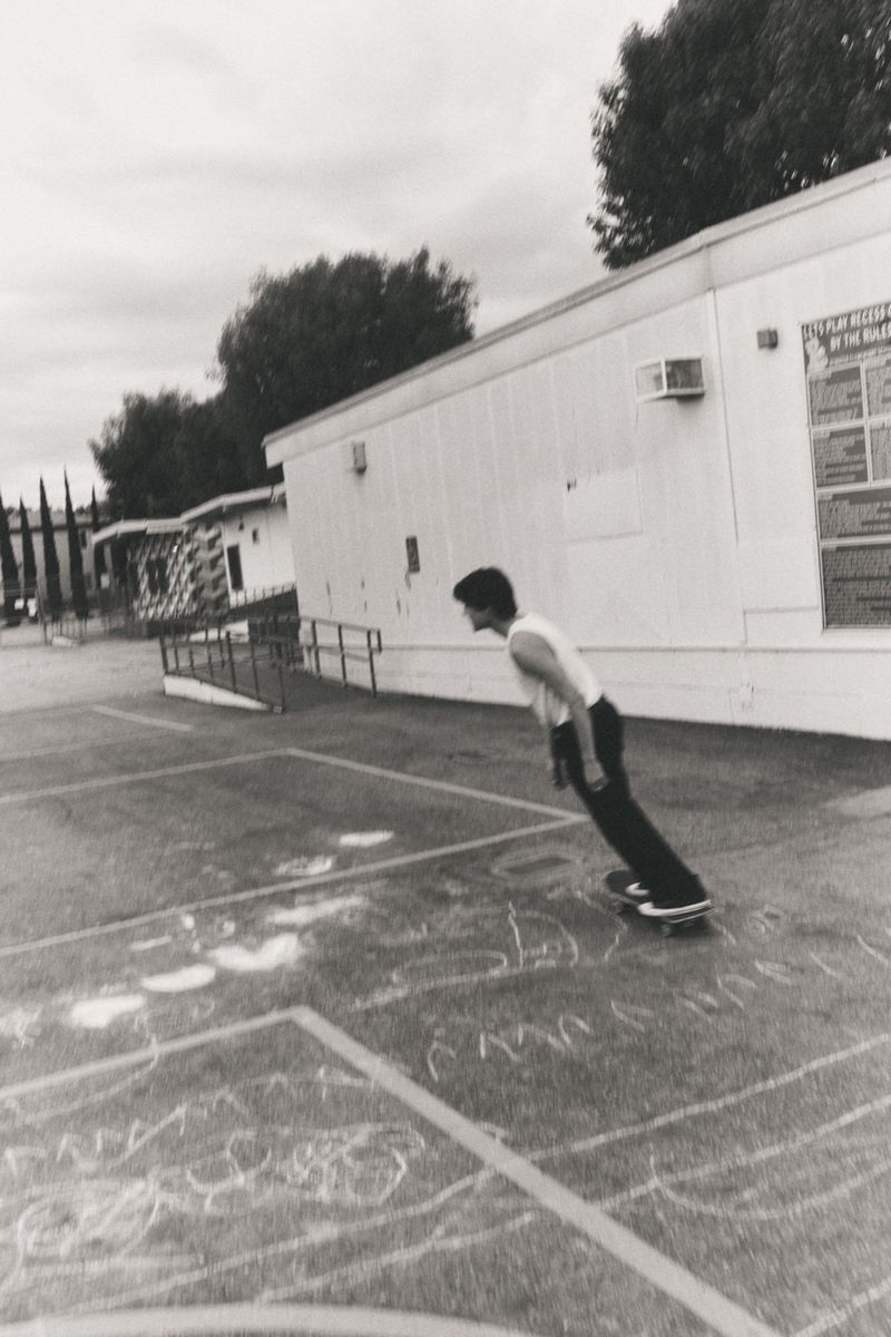 Monochrome image captures a young adult on a skateboard in an urban outdoor setting, with a school building in the background and cloudy sky.