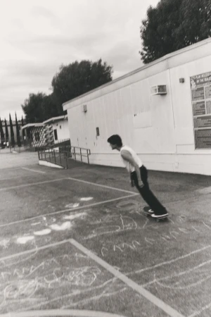 Monochrome image captures a young adult on a skateboard in an urban outdoor setting, with a school building in the background and cloudy sky.