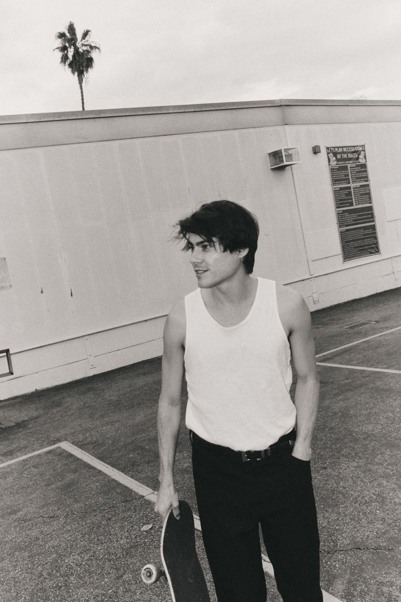 Black and white shot of a young man in a tank top and black pants holding a skateboard, standing in front of a light-colored building.