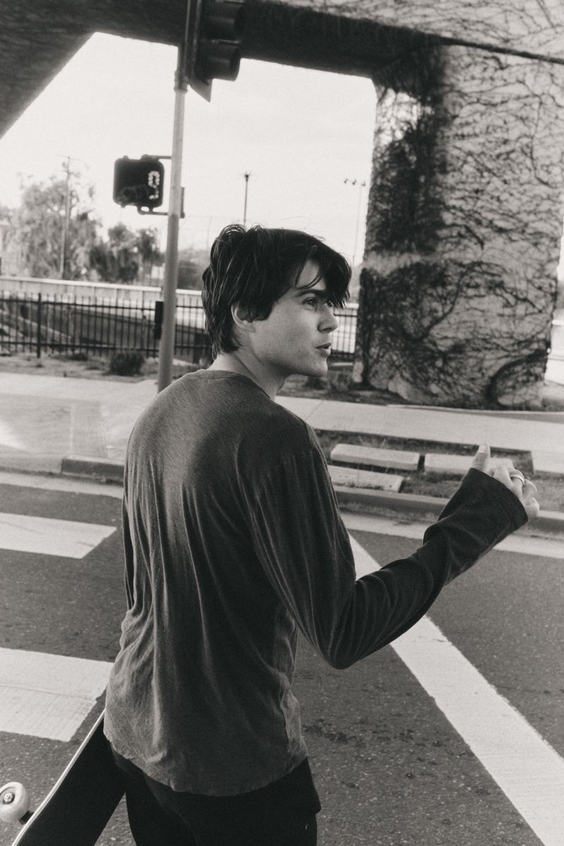 Monochrome shot of a young adult male walking across a crosswalk with a skateboard, captured in an urban environment with an overpass.