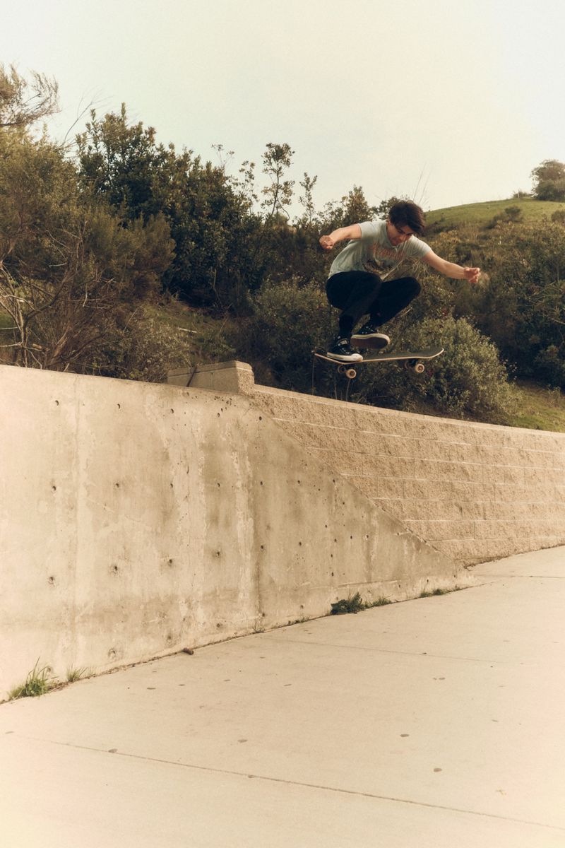 Teen male skateboarding in mid-air against a concrete wall and grassy hill backdrop, capturing an action moment with an outdoor setting.