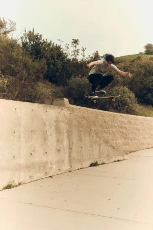 Teen male skateboarding in mid-air against a concrete wall and grassy hill backdrop, capturing an action moment with an outdoor setting.