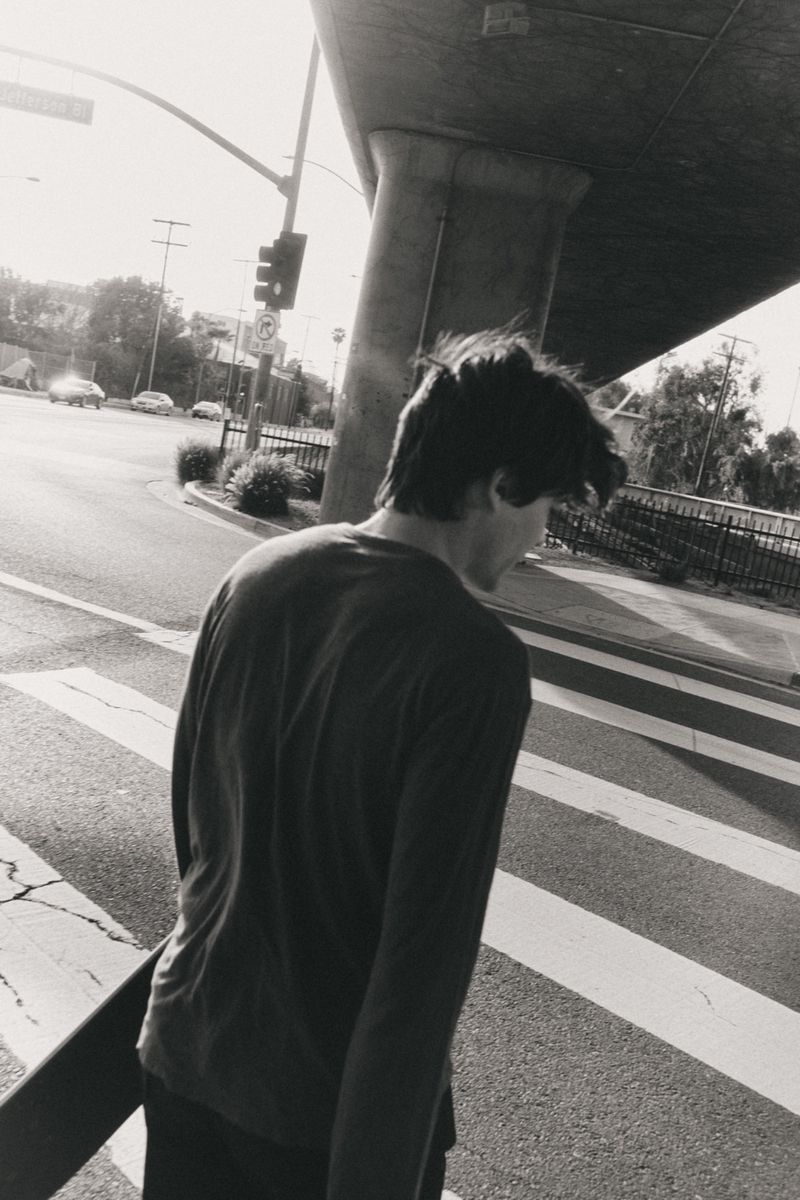 Black and white street photography featuring a young man walking across a crosswalk carrying a skateboard in an urban environment.