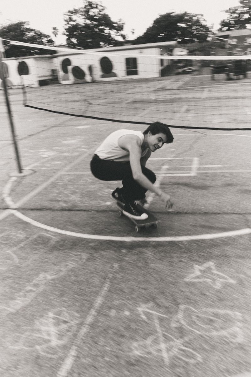 Monochrome image of a young adult skateboarding on a concrete court marked with chalk lines, in an outdoor setting with a building and trees.