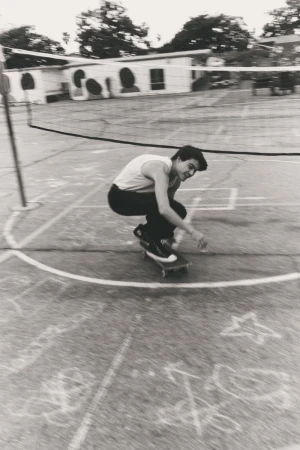 Monochrome image of a young adult skateboarding on a concrete court marked with chalk lines, in an outdoor setting with a building and trees.