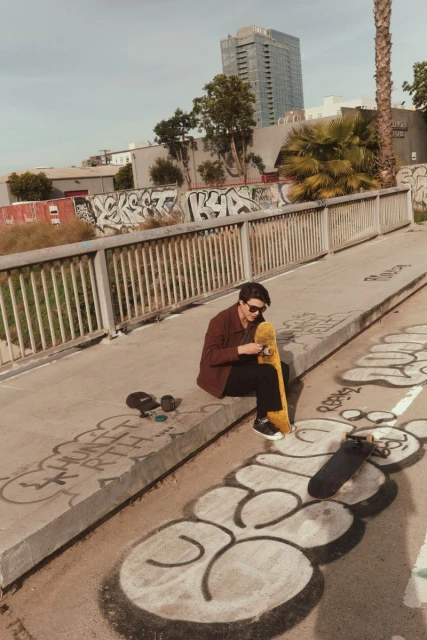 A young man in a burgundy jacket sits on a concrete ledge next to two skateboards with a yellow skateboard in his lap, urban environment.
