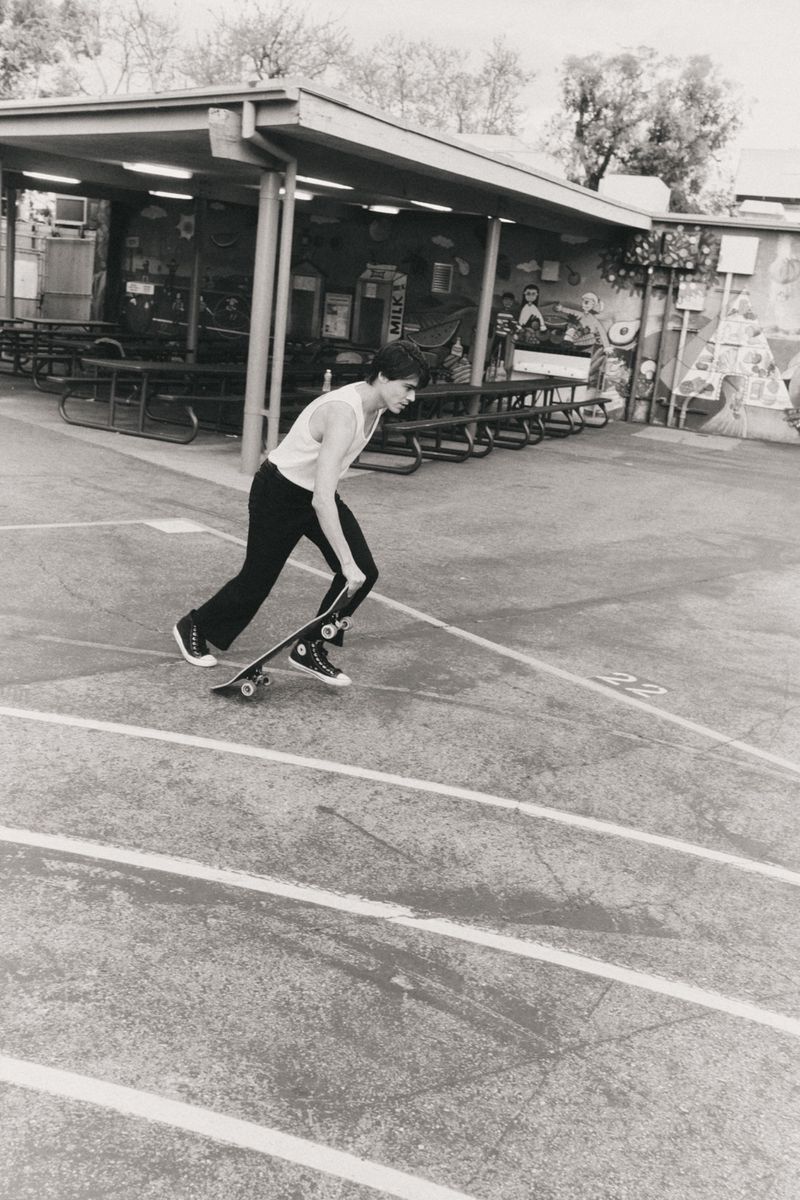 Black and white shot of a young man skateboarding on a marked course outside, next to an outdoor covered eating area.