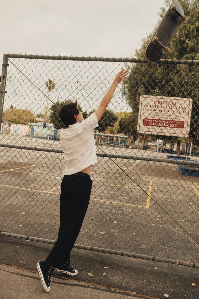 Teenager in white shirt and black pants reaches for a skateboard in the air in front of a chain-link fence outdoors on a sunny day.