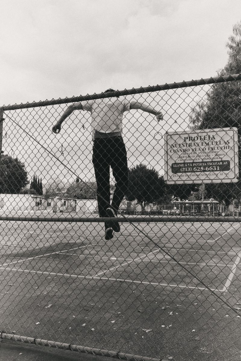 Monochrome photo of a person jumping over a chain-link fence in an outdoor setting with a sign and trees in the background under a cloudy sky.