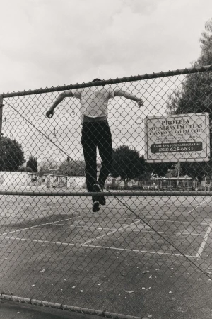 Monochrome photo of a person jumping over a chain-link fence in an outdoor setting with a sign and trees in the background under a cloudy sky.