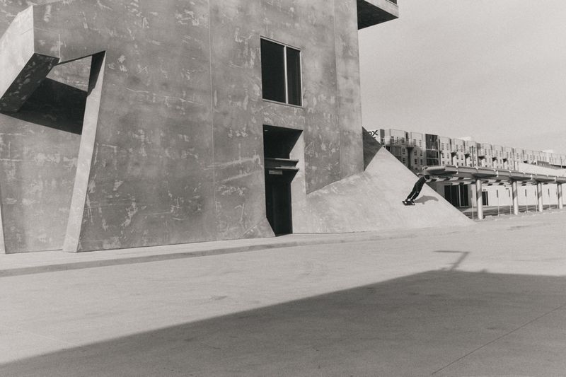 Monochrome image captures a skateboarder gliding down a concrete structure with modern buildings in the background, highlighting urban sport.