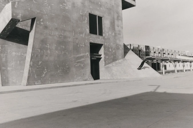 Monochrome image captures a skateboarder gliding down a concrete structure with modern buildings in the background, highlighting urban sport.