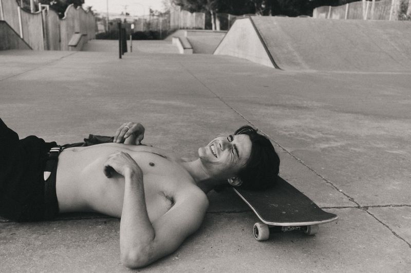 Monochrome shot of shirtless man lying on skateboard in skatepark smiling. Black pants, concrete ramps and walls in background.
