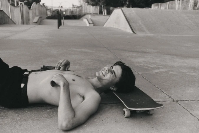 Monochrome shot of shirtless man lying on skateboard in skatepark smiling. Black pants, concrete ramps and walls in background.