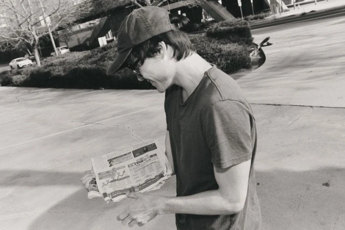 Monochrome outdoor shot of adult male reading newspaper in sunlight, wearing cap, short-sleeve shirt, glasses on concrete surface.