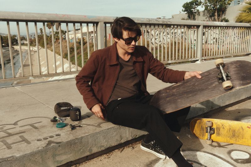 A young man sits on a concrete wall holding a skateboard, dressed in a brown jacket, with another skateboard in the foreground.