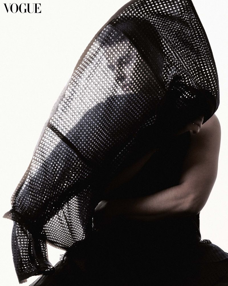 Fashion editorial shot featuring an adult in black headwear with a net pattern, studio lighting, and Vogue magazine logo. 