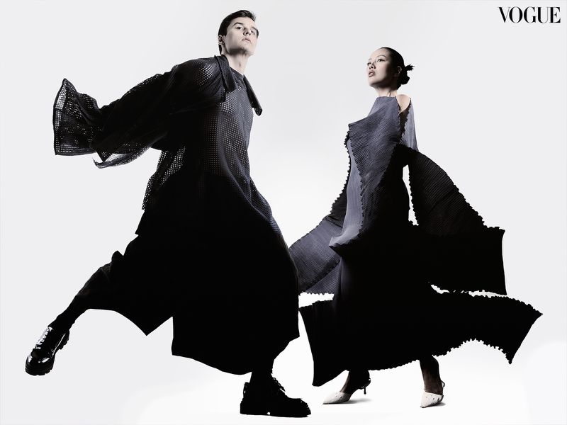 Editorial fashion shot of a male and female model in draped black clothing against a white background, Vogue logo visible.