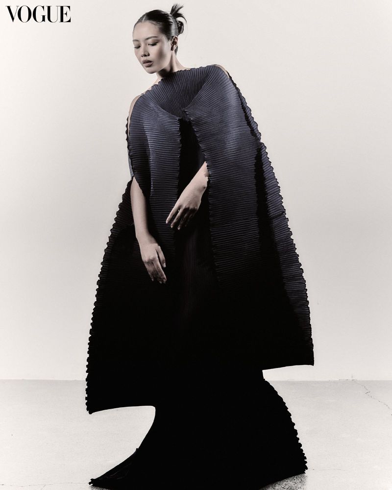 A stylish Asian female model poses gracefully in a striking black pleated dress against a plain background in a fashion editorial shot.