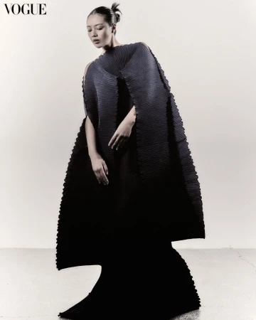 A stylish Asian female model poses gracefully in a striking black pleated dress against a plain background in a fashion editorial shot.