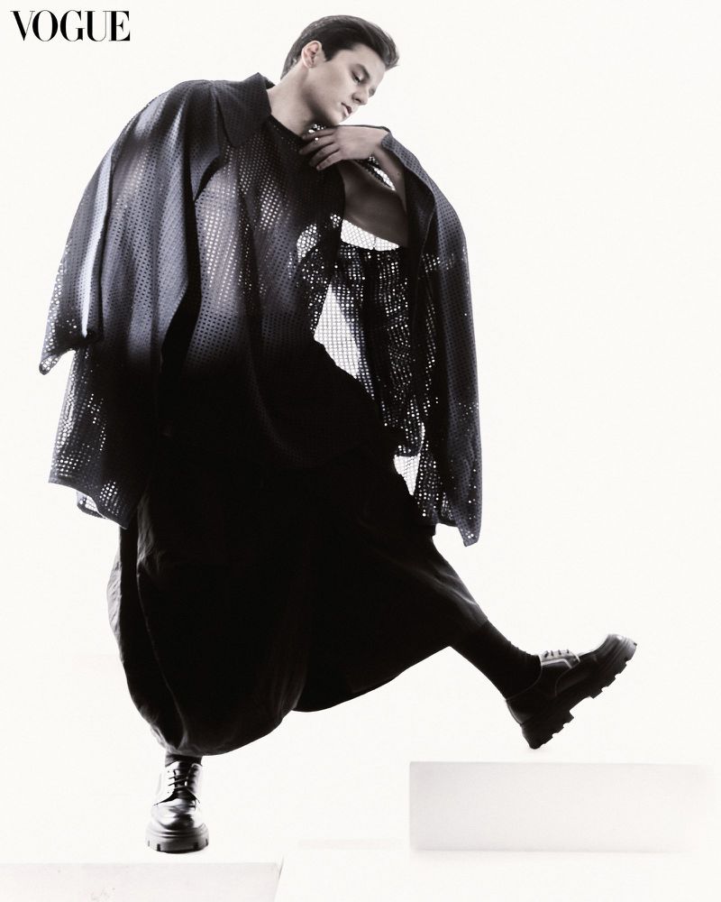 Fashion shot of a young adult male model in a black, see-through poncho and black clothing stepping on a white block in a photo studio.