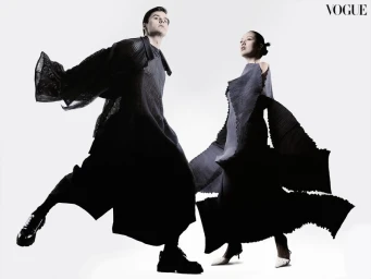 Two models wearing dark flowing pleated outfits posing dynamically against a plain white studio background.