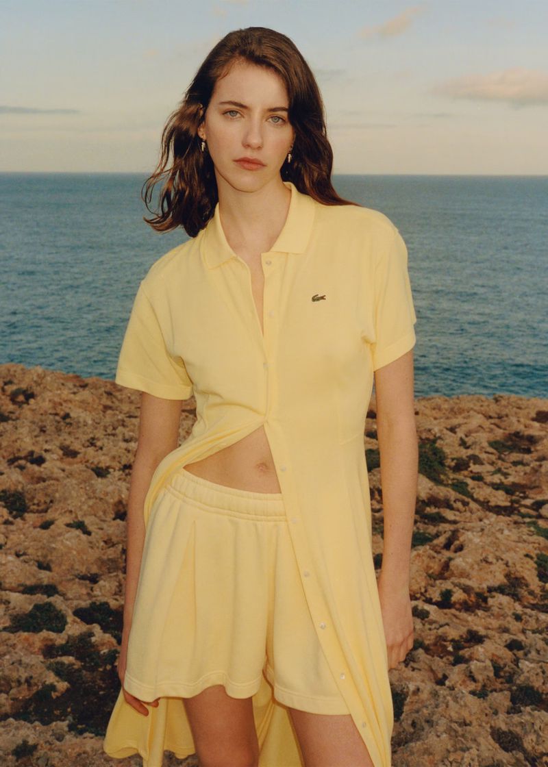 Woman poses in yellow Lacoste polo dress and shorts on rocky coast with ocean backdrop, highlighted by soft natural light. Fashion Editorial.