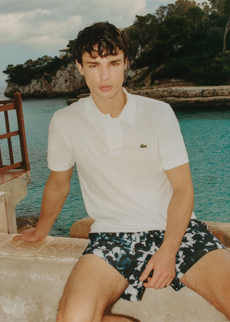 A young, dark-haired man poses, wearing a white polo shirt and printed shorts, sitting outside against a coastal backdrop on a bright day.