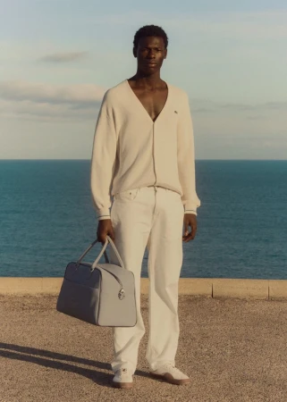 Full shot of male model in casual wear holding a blue duffel bag against a coastal backdrop with a blue ocean and cloudy sky.