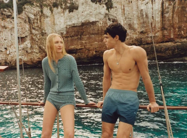 Fashion shoot on a boat featuring a blond woman in knitwear and a shirtless man in swim trunks, with a background of rocky cliffs.