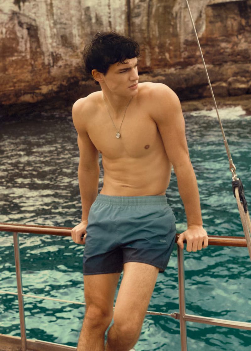 A young adult man in swim trunks poses shirtless on a boat with a rocky cliff and turquoise ocean in the background, looking off to the side.