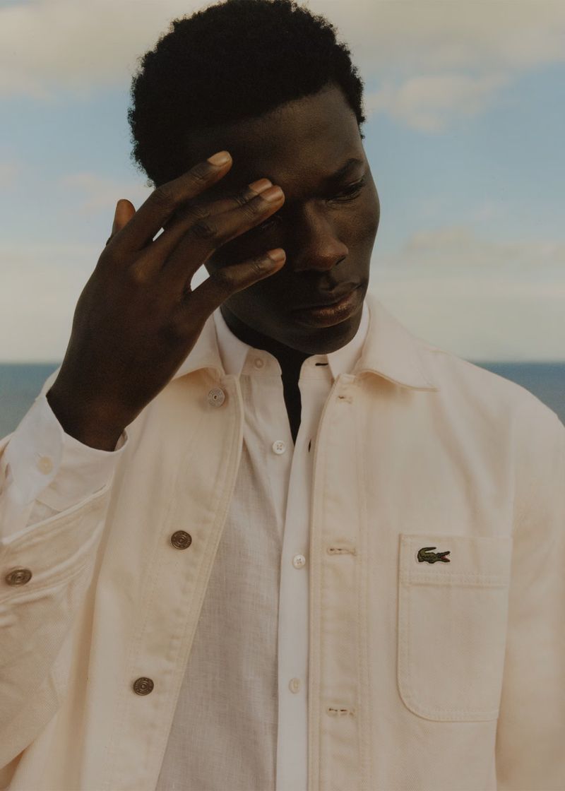 Portrait of a dark-skinned male model in an unbuttoned off-white jacket and white button-down shirt, posing outdoors against a bright sky.