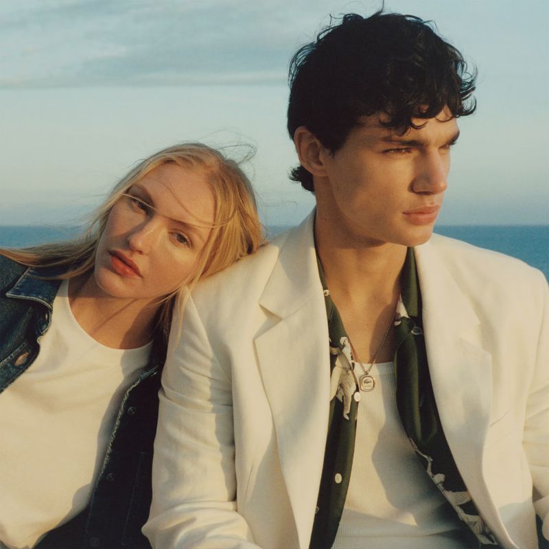 Stylish couple poses outdoors with a backdrop of the ocean and sky. A blonde woman rests her head on a man's shoulder.