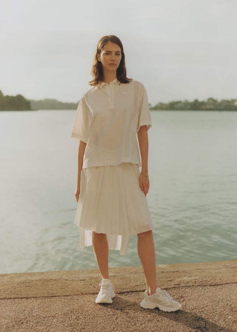 Woman in white blouse and skirt poses by tranquil lake, showcasing fashion apparel in outdoor setting with soft natural light.