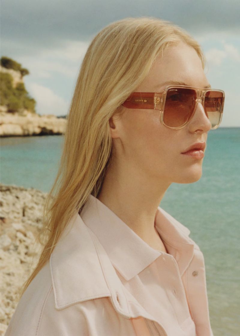 Close-up of a blonde woman in pink jacket and tinted sunglasses posing on a beach with turquoise water and cloudy sky in the background.
