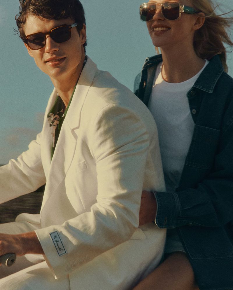Stylish couple wearing sunglasses riding a scooter, the man in a white suit, and the woman in denim, enjoying a sunny day outdoors.