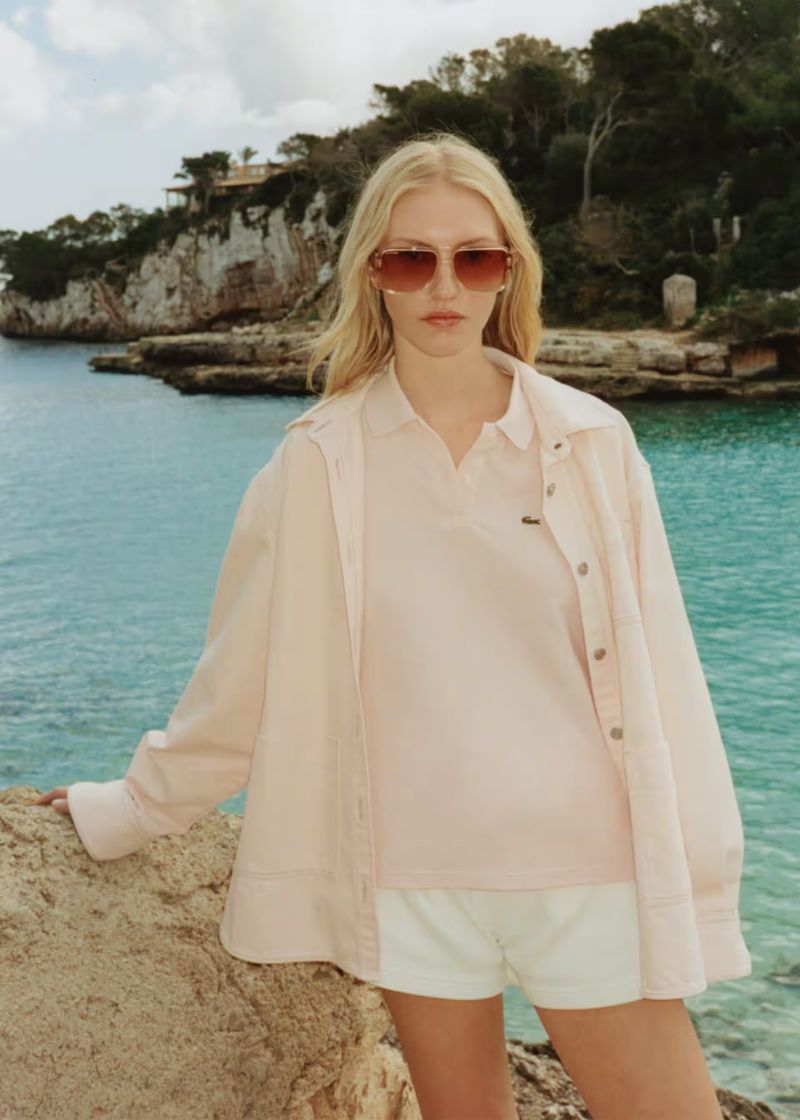A blonde adult woman wearing sunglasses, a pink polo shirt, white shorts, and a pink jacket standing on rocks near a blue ocean.